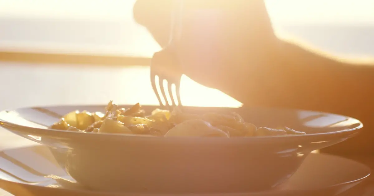 Close-up of a plate of pasta being eaten by the sea on the beach, with a sunset in the background.