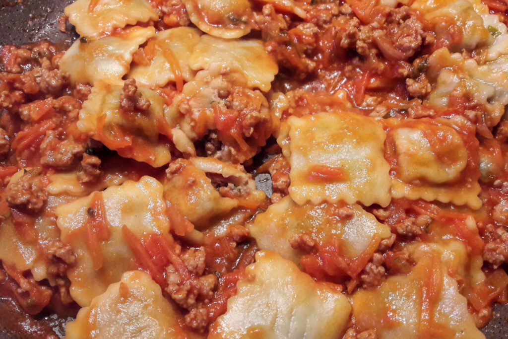 A close-up image of some tordelli with ragu, a square-shaped filled pasta with serrated edges, dipped in a rich tomato sauce with minced meat.