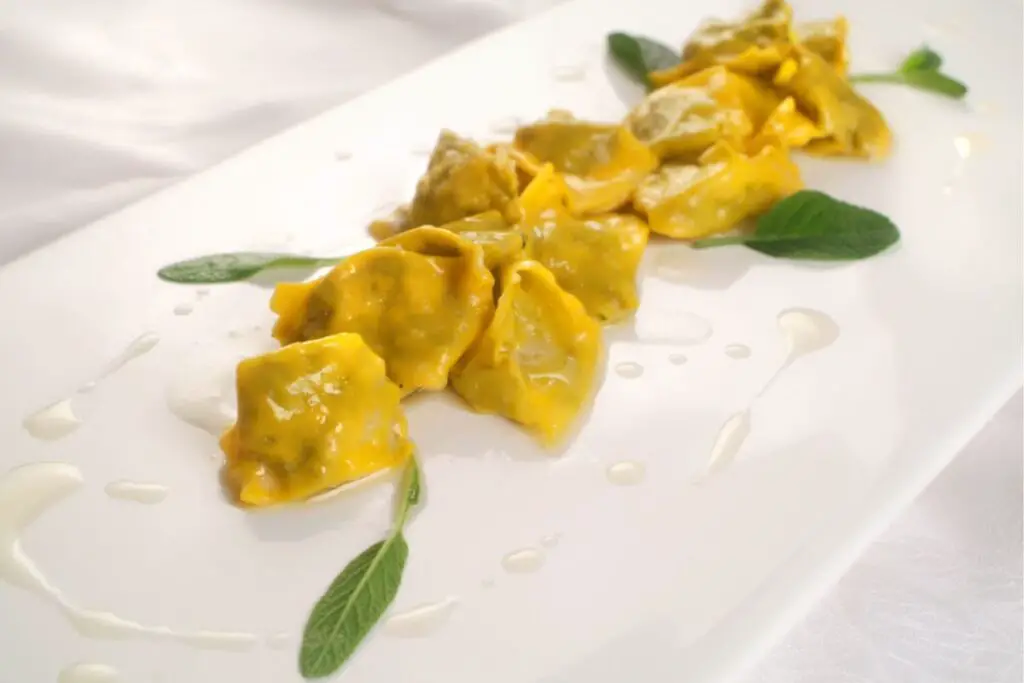 An oblong plate with agnolotti arranged in a row on top, a type of filled pasta with an irregular and squat shape. They are bathed in a transparent sauce of melted butter and flavored with sage leaves.
