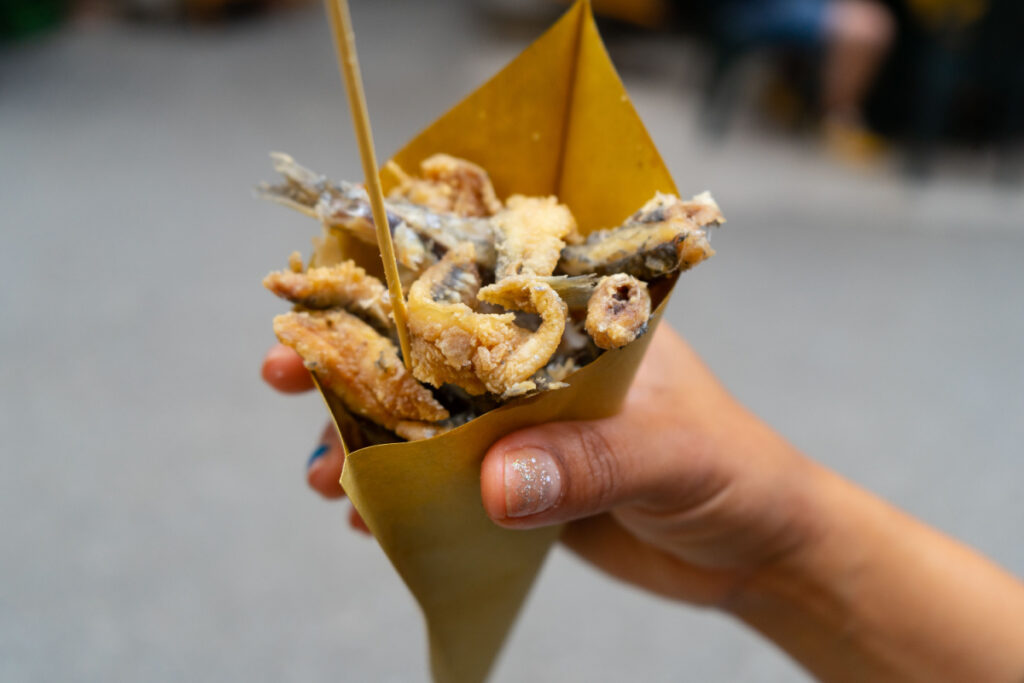 A hand holds a paper cone full of fried fish, squid and small fish can be seen coming out of the cone. All golden and crunchy battered.