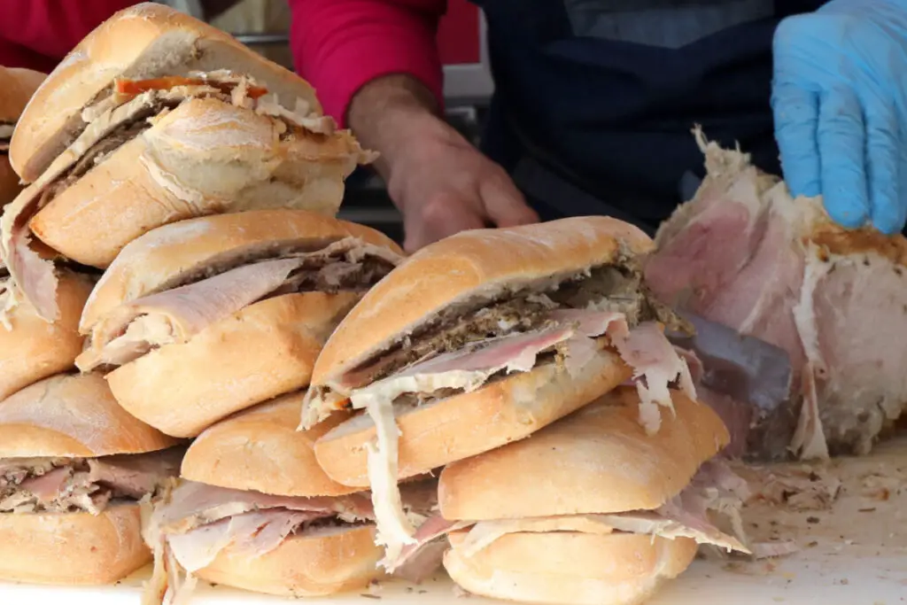 A series of porchetta sandwiches piled up on the butcher's counter. Nearby the butcher is slicing pieces of porchetta with which to stuff other sandwiches.