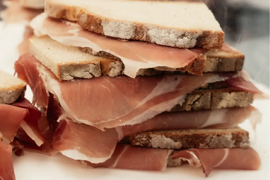 A pile of sandwiches made with thin slice of bread filled with prosciutto cured meat.