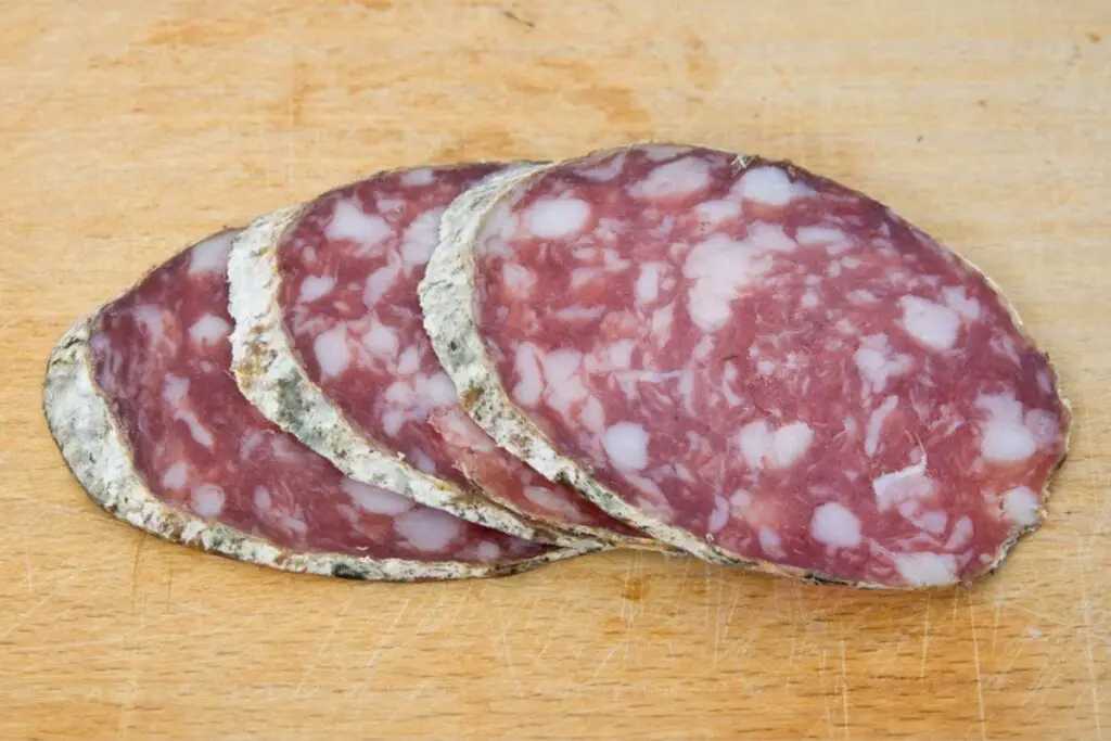 Slices of Cinta Senese salami placed on a wooden cutting board are thick and the meat is darker than a normal salami.