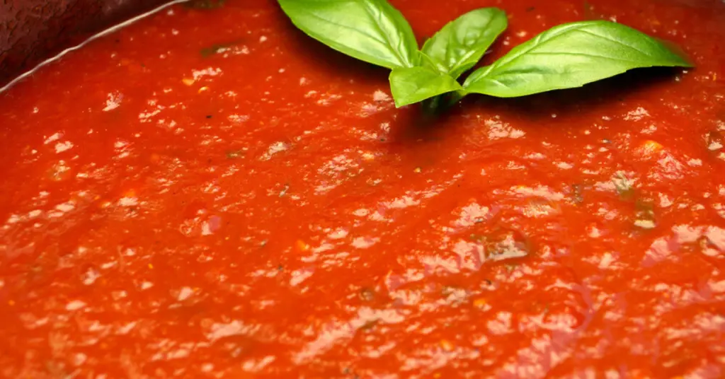 A close-up photo of marinara sauce, with pieces of basil and oregano visible in the red tomato sauce.