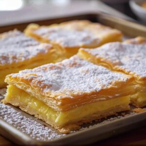 Rectangles of very light puff pastry that enclose a heart of custard, are dusted with icing sugar and are placed on a tray on the top of a home kitchen.