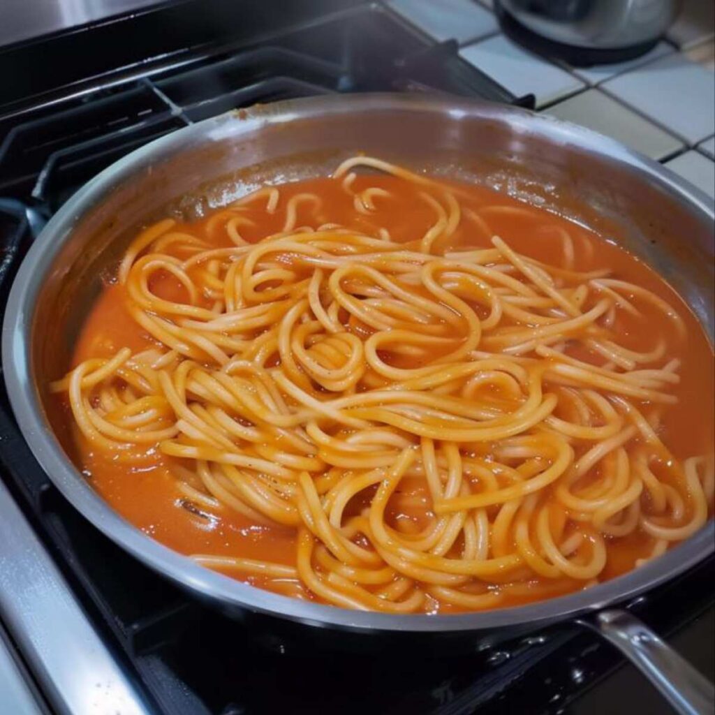 A pan on the stove, inside it, pici are cooking, a type of pasta that resembles very thick spaghetti, along with a tomato and garlic sauce, with a reddish-orange color, the sauce is thickening and simmering.