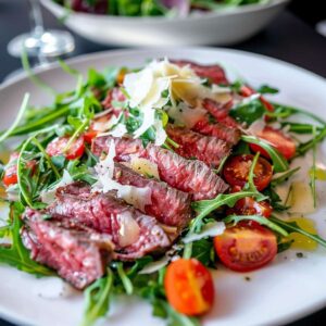 Dish with sliced jiuce beef steak served with arugula and sliced cherry tomatoes, topped with Parmesan shavings.
