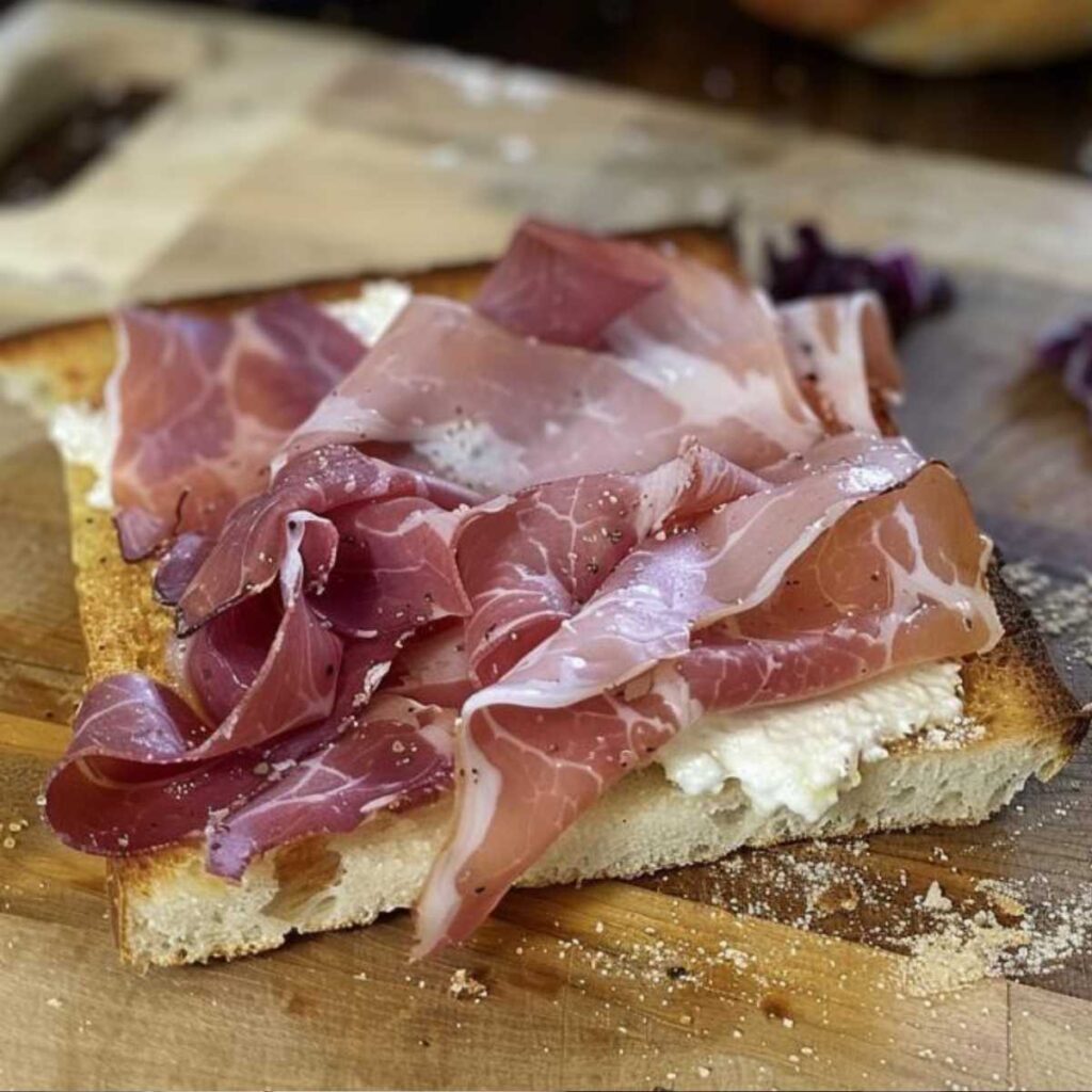The image shows a Tuscan schiacciata, a type of flat, rustic bread, sliced and filled with toppings. The top of the bread is slightly curved, suggesting a crunchy crust with a porous interior. The filling includes a lush layer of red wine radicchio cream, a soft mixture with a deep, dark purple color, indicative of a slow cooking process that enhances its flavors. Above the radicchio cream is a generous amount of gorgonzola cream, which appears smooth and easily spreadable, with its characteristic creamy white hue marbled with green-blue veins. Topping off this creation are translucent slices of speck, a cured meat with colors ranging from pale pink to dark red, a sign of proper curing and smoking. The slices are enticingly arranged, folded slightly to add volume and depth to the filling. The hands holding the schiacciata appear ready to enjoy this delicious and hearty morsel