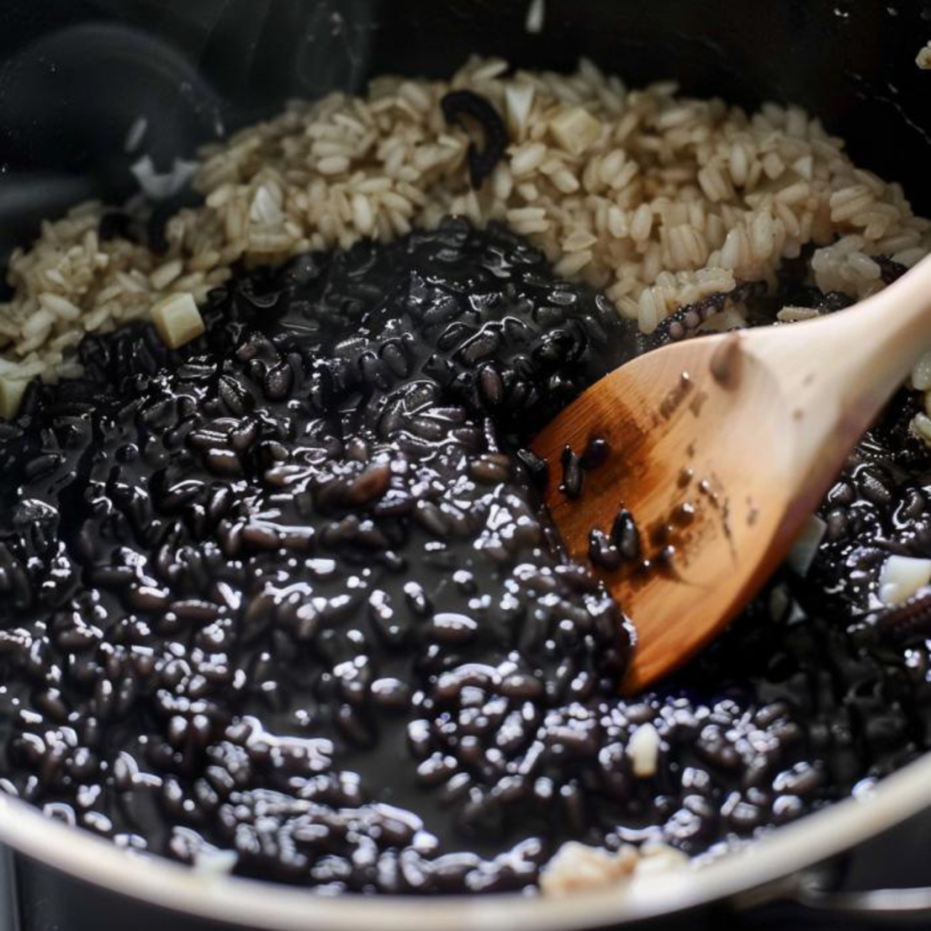 In this photo, there is a wide pot containing squid ink risotto being stirred with a wooden spoon. The risotto shows a variety of shades: the part submerged in the squid ink is intensely black and glossy, while some grains on the surface remain white, indicating that the dish is still cooking and the ink has not yet uniformly colored all the rice. Among the rice, there are visible pieces of squid, likely cut into cubes, which add texture and flavor to the dish. The presence of the squid pieces and the contrast between the white and black rice create a captivating visual effect.