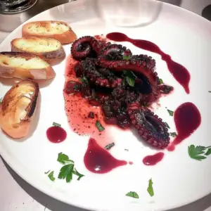 This dish features octopus, cooked and plated with a modern aesthetic. The octopus has a dark hue, indicative of being stewed in red wine, a technique used to create the "drunken" effect. Accompanying the octopus are symmetrical toast points along the plate’s edges, adding crunch and acting as a sponge for the savory sauce. At the center, the octopus is prominently displayed with whole tentacles and cut pieces revealing a tender texture. The red wine sauce pools around the octopus, bringing life and color to the dish. Scattered chopped parsley on the side of the plate adds a pop of fresh color and a nod to the aromatic herbs typical of Mediterranean cuisine.