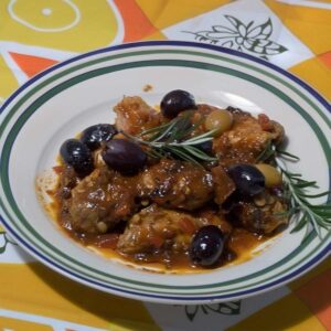 The picture presents a hearty portion of rabbit cacciatore, its pieces generously coated in a thick, glossy sauce that hints at a slow, savory cooking process. The olives, a mix of black and dark purple hues, not only add a visual contrast but also suggest a briny, rich accent to the dish's flavor profile. The plate itself is classic, with a clean white center bordered by concentric circles of green and navy, suggesting a traditional or possibly vintage style. This carefully plated dish is set upon a surface featuring a sunny, geometric pattern--a yellow floral design against a backdrop of alternating orange and cream stripes, all of which lend a distinctly retro and cheerful ambiance to the meal's presentation. The setting is casual yet thoughtful, evocative of a homey and inviting dining experience.
