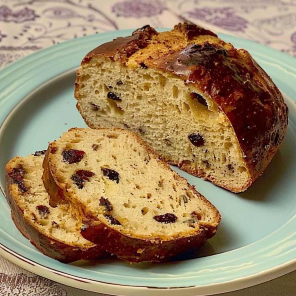 The image displays a whole and sliced Buccellato di Lucca bread placed on a pale blue plate. The bread features a well-baked crust with dark, crunchy areas. The interior is beige in color, with a texture that appears soft and porous, and it's generously studded with raisins. 