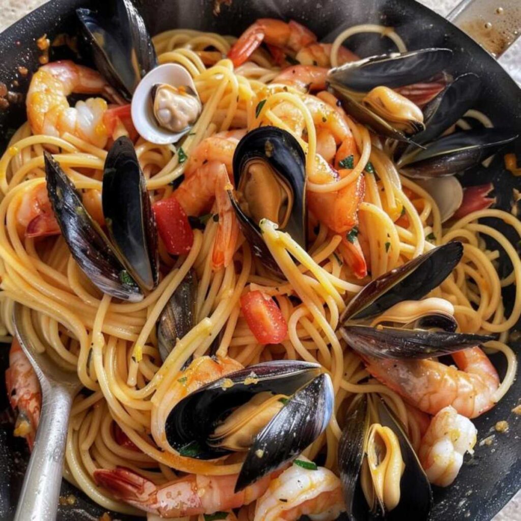 This image presents a close-up view of a pan full of spaghetti allo scoglio, a traditional Italian seafood pasta dish. The spaghetti is cooked and appears to be tossed with a light sauce. Among the pasta are a variety of seafood: open mussels with their blue-black shells, whole shrimps with their orange-pink shells, and clams with their striped, pale shells. Pieces of tomato and herbs are scattered throughout, adding color and suggesting a flavorful, aromatic experience. The dish is a feast for the eyes, with the vibrant colors and textures suggesting a fresh and tasty meal straight from the sea.
