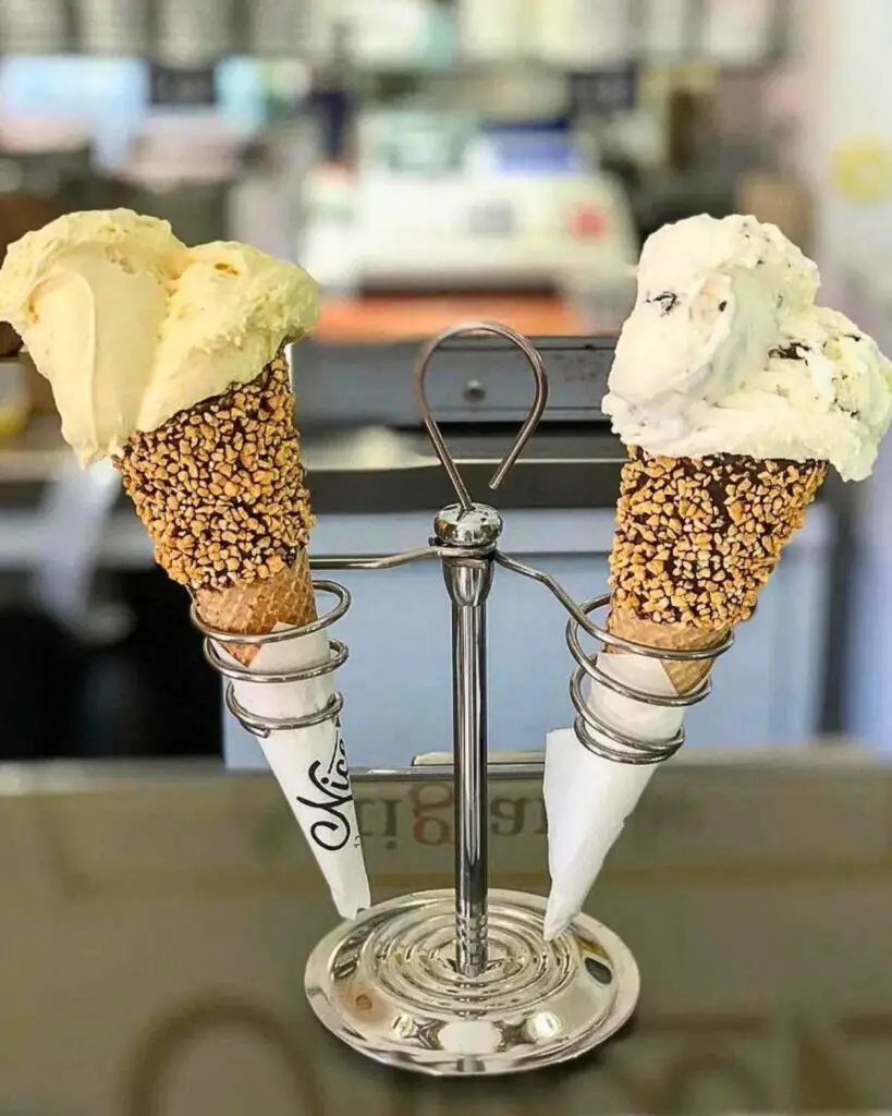 Two gelato cones topped with delicious scoops, one vanilla and one chocolate chip, served in crunchy, nut-covered cones at Gelateria Nice.