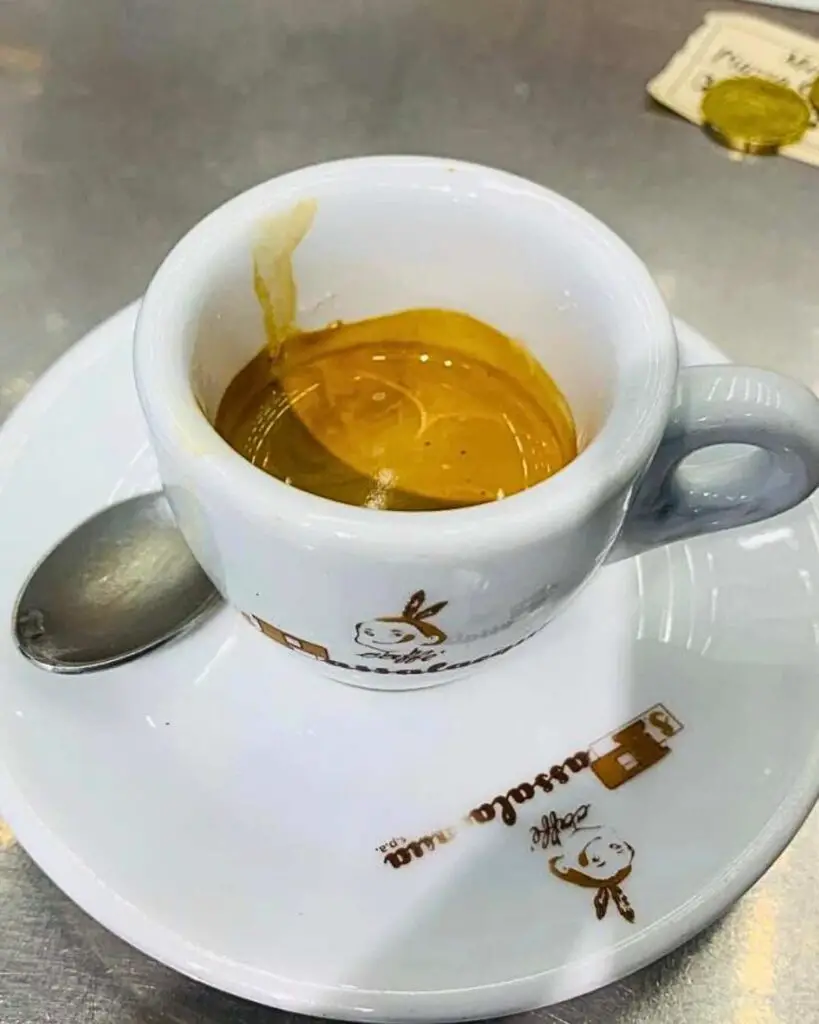 A freshly made espresso served in a white cup with a brown logo, placed on a white saucer with a small spoon, showcasing the traditional coffee in Naples Italy at Bar Mexico, one of the best cafes in Naples.