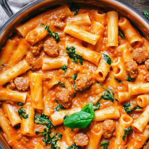 A close-up view of creamy sausage rigatoni pasta, featuring rigatoni pasta cooked in a rich tomato-based sauce with Italian sausages and fresh spinach. The dish is presented in a stainless steel skillet with the sauce clinging to the rigatoni, highlighting the ridges of the pasta. Fresh herbs are scattered around, adding a pop of color and flavor. The golden-brown sausage bits add texture, while the creamy sauce provides a luxurious finish, making it a hearty and comforting Italian meal.