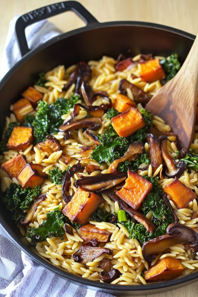 A skillet filled with fall orzo pasta, caramelized mushrooms, roasted sweet potatoes, sautéed kale, and onions, showcasing a colorful blend of textures and flavors.