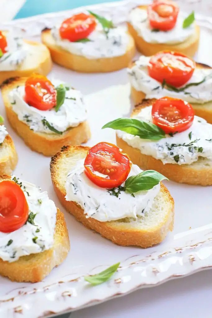 Crostini topped with a creamy herb cream cheese spread, sliced grape tomatoes, and fresh basil, served on toasted baguette slices as a light tomato appetizer