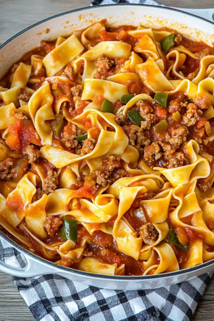 A savory dish of Italian drunken noodles featuring wide, ribbon-like pasta combined with crumbled Italian sausage, cooked in a rich tomato-based sauce with bell peppers and onions. The dish is served in a large skillet, perfect for sharing, and offers a flavorful fusion of Italian and Asian influences.