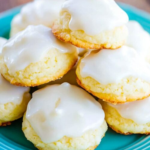 A stack of traditional Italian lemon drop Christmas cookies on a blue plate, covered in a thick lemon glaze. The cookies are soft and golden, with a subtle biscuit-like texture, offering a burst of citrus flavor from the fresh lemon glaze. Perfect for anyone seeking authentic Italian Christmas cookies recipes, these treats are a holiday favorite.