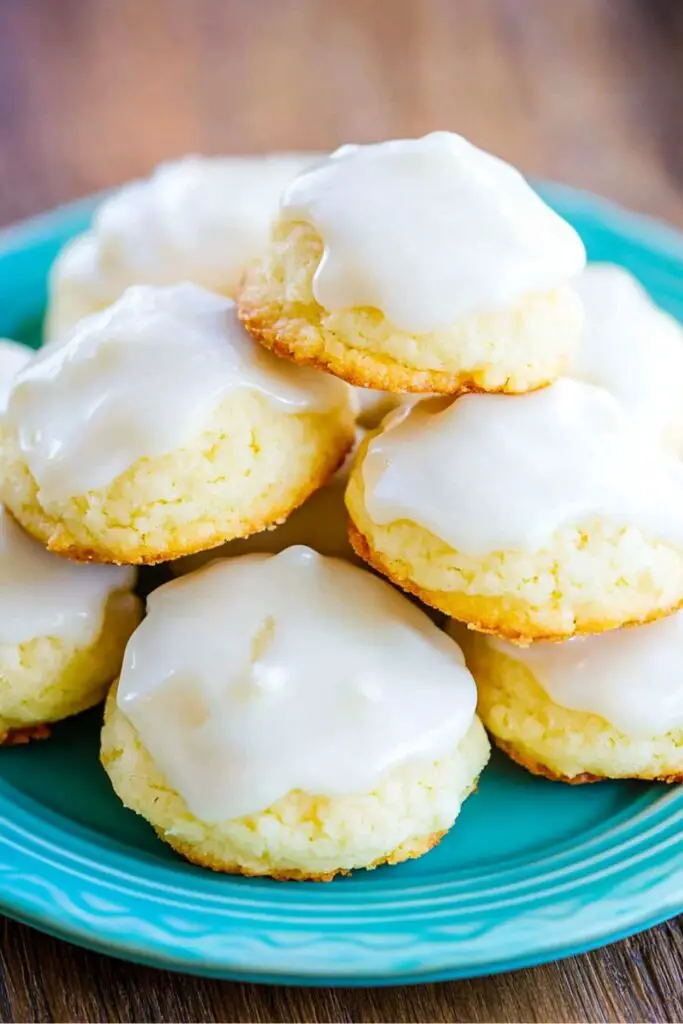 A stack of traditional Italian lemon drop Christmas cookies on a blue plate, covered in a thick lemon glaze. The cookies are soft and golden, with a subtle biscuit-like texture, offering a burst of citrus flavor from the fresh lemon glaze. Perfect for anyone seeking authentic Italian Christmas cookies recipes, these treats are a holiday favorite.