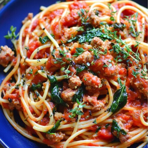 A vibrant close-up of Italian sausage spaghetti served in a blue bowl. The spaghetti is coated in a rich tomato marinara sauce, mixed with crumbled Italian sausage, fresh spinach, and diced tomatoes. The dish is garnished with fresh thyme, adding a touch of greenery and a burst of flavor to the meal. This hearty pasta is a perfect blend of flavors, showcasing Italian sausage as the star ingredient.