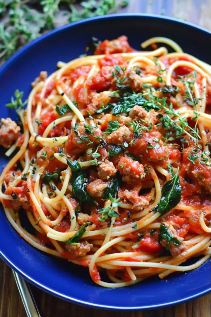 A vibrant close-up of Italian sausage spaghetti served in a blue bowl. The spaghetti is coated in a rich tomato marinara sauce, mixed with crumbled Italian sausage, fresh spinach, and diced tomatoes. The dish is garnished with fresh thyme, adding a touch of greenery and a burst of flavor to the meal. This hearty pasta is a perfect blend of flavors, showcasing Italian sausage as the star ingredient.