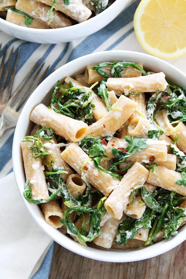 A bowl of rigatoni pasta tossed with creamy burrata, fresh arugula, and a zesty lemon sauce, garnished with red pepper flakes and served with a lemon wedge on the side.