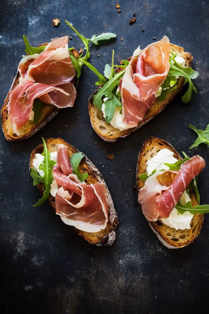 Crispy toasted bruschetta topped with thin slices of prosciutto and shavings of parmesan cheese, drizzled with olive oil, served as a savory Italian appetizer for a cocktail party