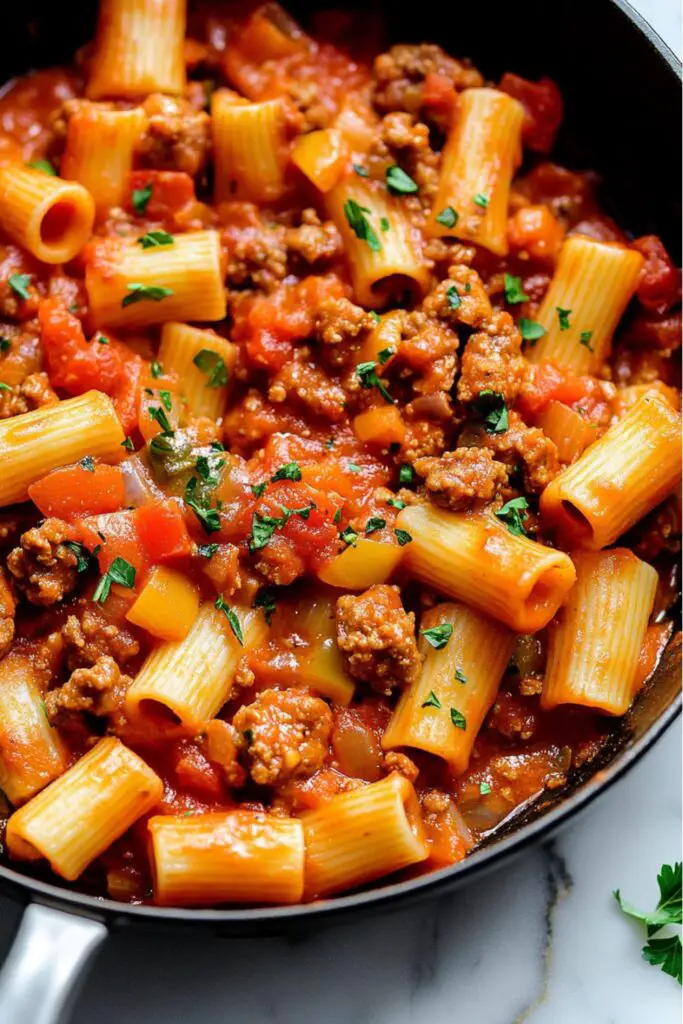 A close-up view of rigatoni pasta in a rich tomato sauce with crumbled spicy Italian sausage and diced sweet bell peppers. The pasta is garnished with fresh parsley and served in a dark skillet, showcasing a vibrant, flavorful mix of ingredients. The hearty dish is a perfect combination of bold, spicy sausage and sweet, juicy peppers in a savory sauce.