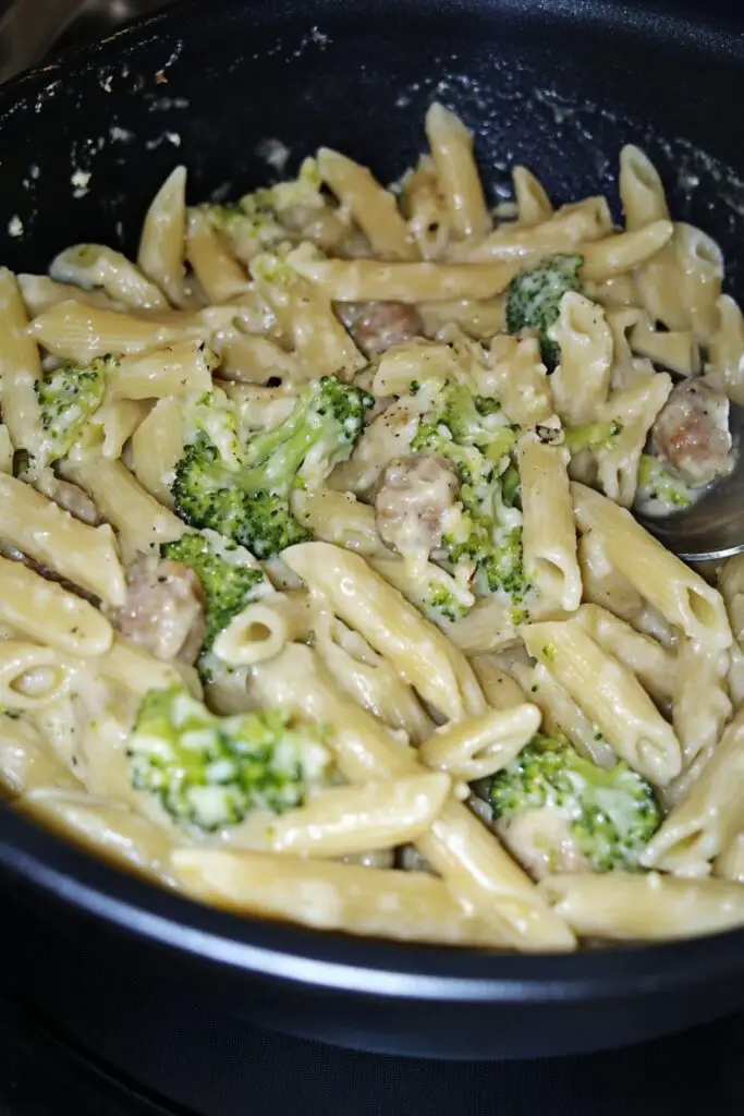 A serving of creamy penne pasta mixed with bite-sized chunks of sweet Italian sausage and broccoli florets. The dish is lightly coated in a garlic-infused sauce with melted cheese and olive oil, served in a dark skillet. The rich, savory flavors of the sausage blend perfectly with the tender broccoli and pasta.