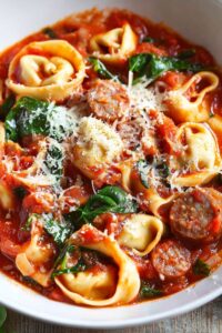 A close-up image of Tuscan tortellini pasta with Italian sausage, spinach, and a rich tomato-based sauce, served in a white bowl. The dish is topped with shredded Parmesan cheese and garnished with fresh herbs. The tortellini is tender, and the sausage bits are mixed into the sauce, creating a hearty and flavorful Italian meal, perfect for busy weeknights.