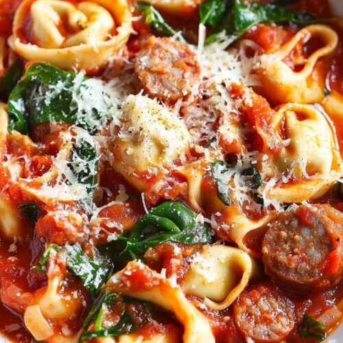 A close-up image of Tuscan tortellini pasta with Italian sausage, spinach, and a rich tomato-based sauce, served in a white bowl. The dish is topped with shredded Parmesan cheese and garnished with fresh herbs. The tortellini is tender, and the sausage bits are mixed into the sauce, creating a hearty and flavorful Italian meal, perfect for busy weeknights.