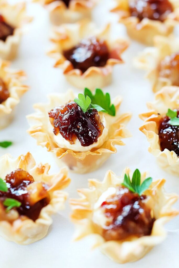 Mini baked brie bites in phyllo cups topped with fig jam and garnished with parsley on a white platter, perfect for a festive Christmas buffet.