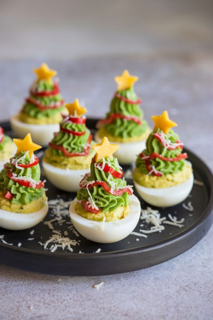 Deviled eggs decorated as miniature Christmas trees with green filling, red pepper "ornaments," Parmesan cheese "snow," and small yellow stars on top, displayed on a tray for a festive holiday appetizer.