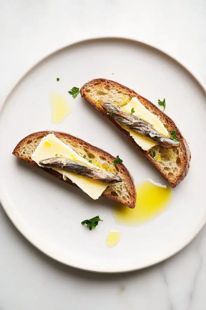 Two crostini topped with butter and marinated anchovies, served on a white plate. A simple yet flavorful appetizer perfect for a traditional Italian Christmas Eve dinner with a focus on seafood dishes.