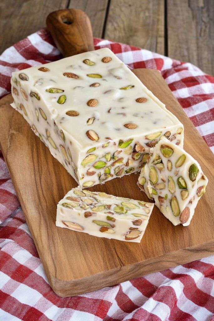 Traditional soft Italian nougat candy with almonds and pistachios, displayed on a wooden board with a red and white checkered cloth in the background.