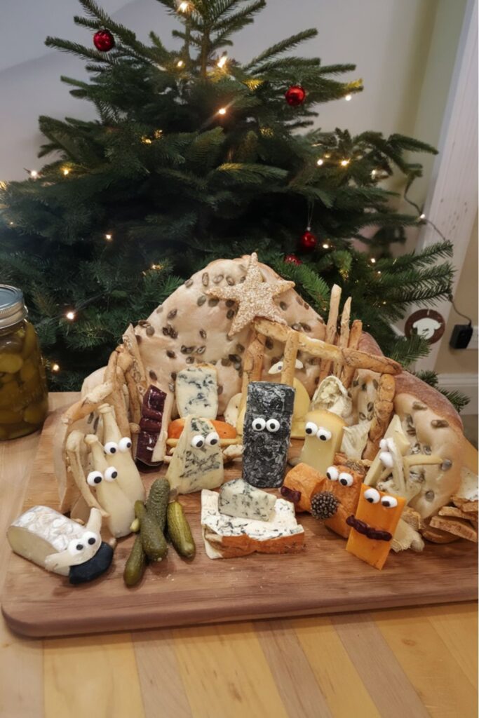 A creative Christmas food charcuterie board shaped like a nativity scene, featuring various cheeses with googly eyes and a backdrop of breadsticks and crackers resembling a manger, surrounded by pickles and olives.