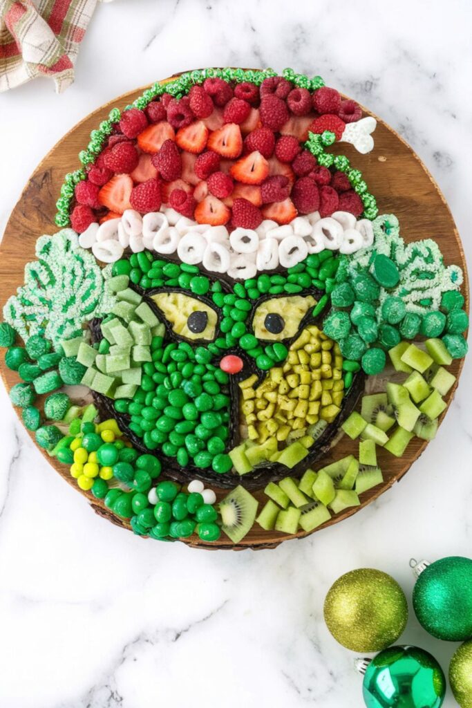 A festive Grinch-themed Christmas dessert charcuterie board featuring green candies, fruits like grapes and kiwi, and chocolate-covered treats arranged to resemble the Grinch's face, with strawberries, raspberries, and pretzels forming his iconic red hat.