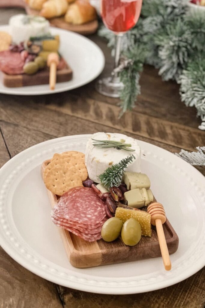 A small Christmas charcuterie board with salami, green olives, creamy cheese topped with rosemary, crackers, honeycomb, and a mini decorative Christmas tree on a wooden plate.