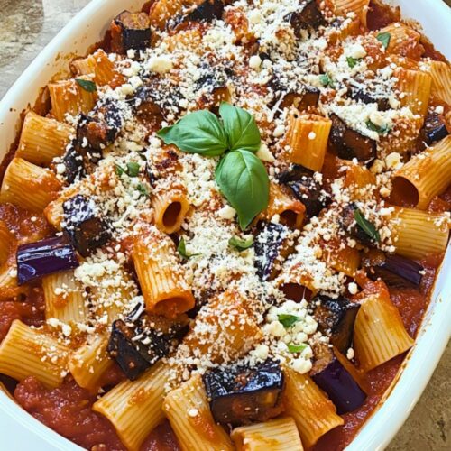 A large white oval dish brimming with pasta alla Norma, showcasing rigatoni in vibrant tomato sauce, caramelized eggplant pieces, a topping of grated ricotta salata, and fresh basil leaves, set on a rustic wooden kitchen table.