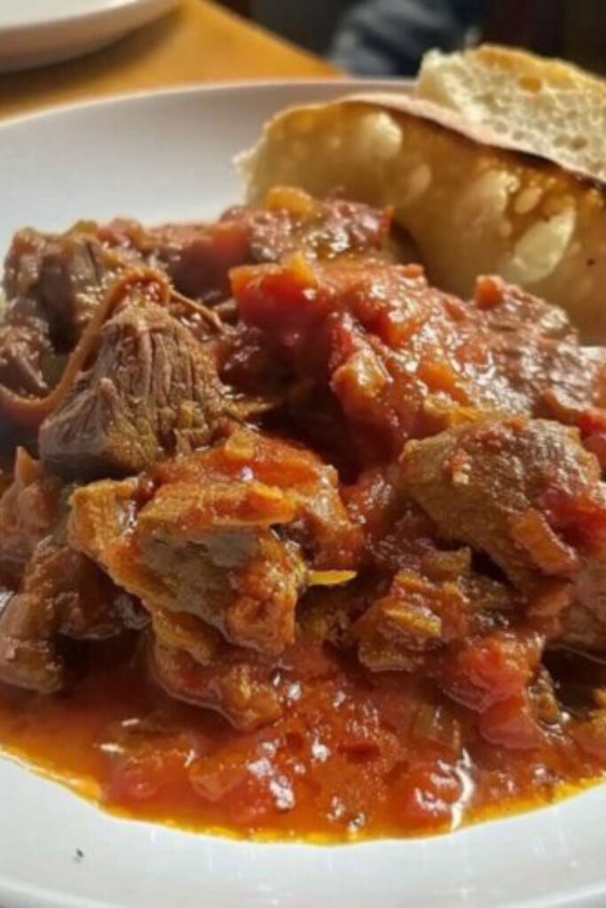 A white bowl filled with a rich tomato-based lamb stew served alongside toasted slices of bread for a selection of foods for easter dinner.