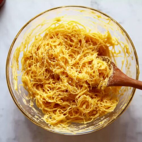 Golden angel hair pasta mixture with Parmesan and pepper in glass bowl on marble countertop.