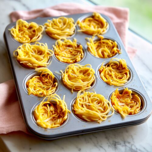 Angel hair pasta nests pressed into a gray metal cupcake pan on a marble countertop.