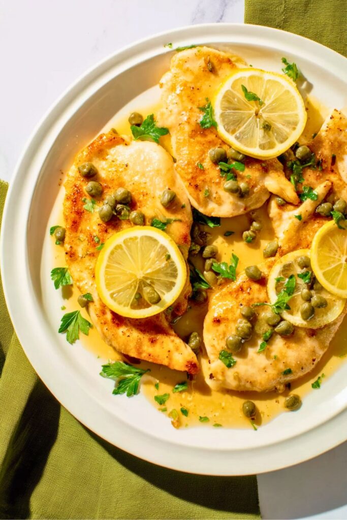 Four golden chicken cutlets topped with lemon slices, capers, and fresh parsley on a white plate.