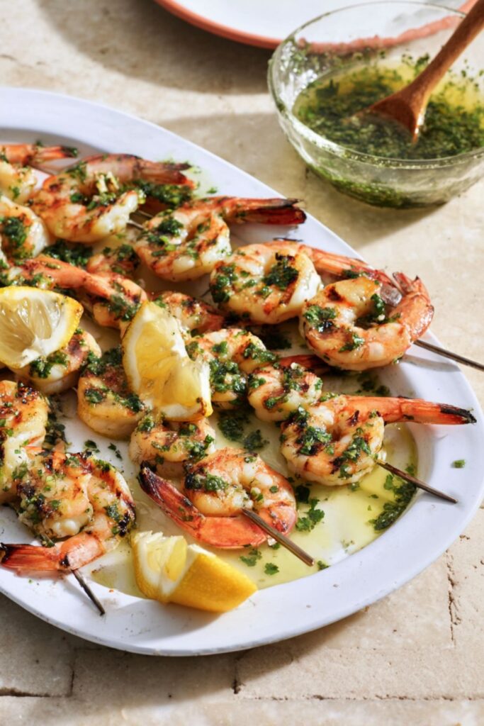 Grilled shrimp skewers topped with fresh salmoriglio sauce, served with lemon wedges on a white platter.
