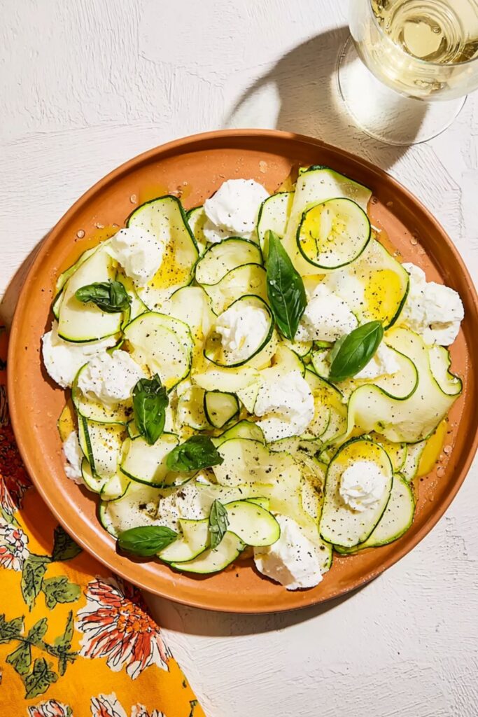 Overhead view of zucchini crudo topped with torn burrata cheese, fresh basil leaves, and lemon zest on a tan plate.