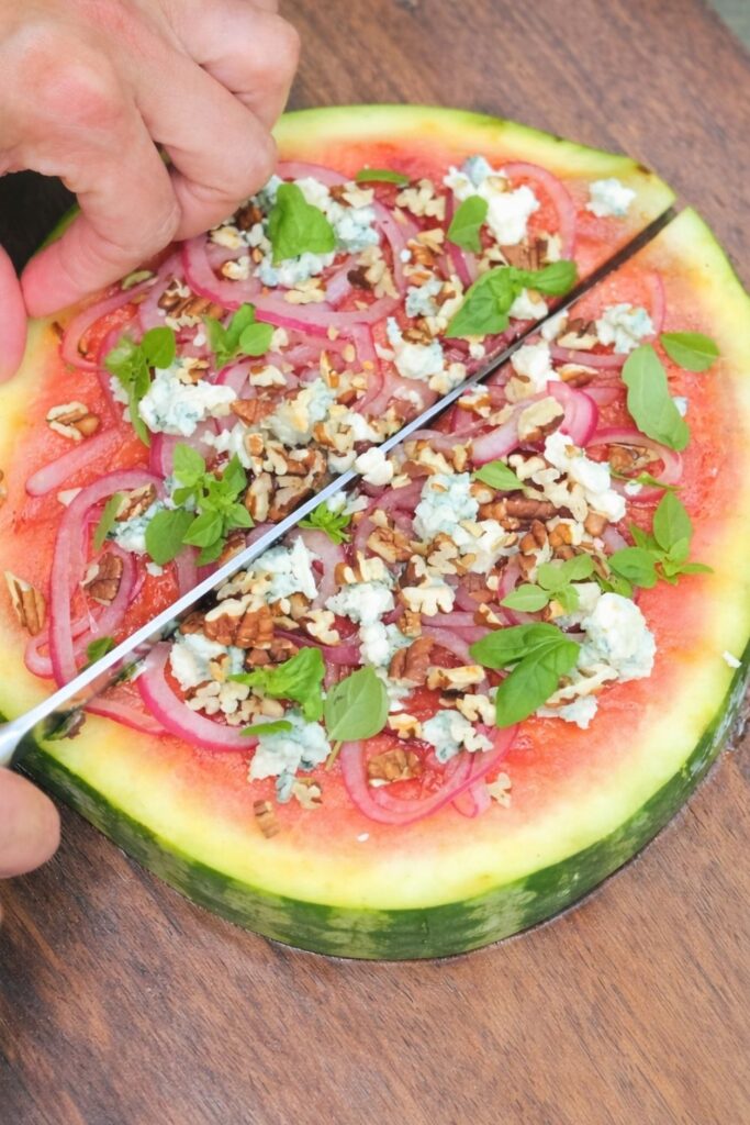 A hand slicing a grilled watermelon pizza topped with onions, cheese, and pecans.