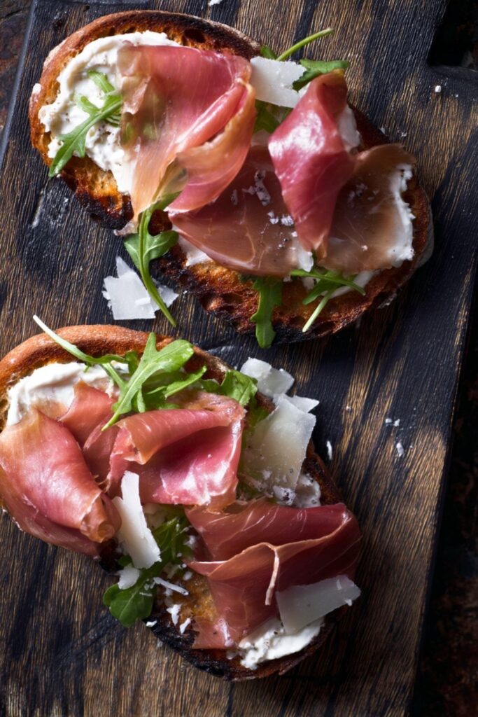 Crispy toasted bread topped with savory prosciutto, cheese, and fresh greens on wood.
