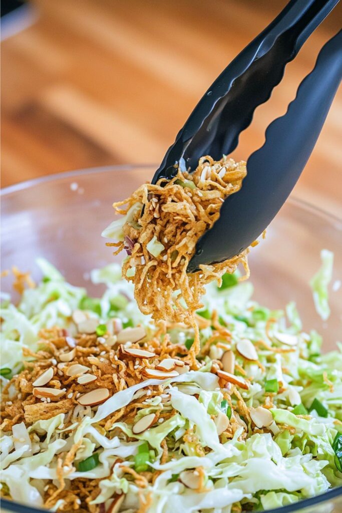 Tongs lifting crunchy ramen and almond salad from a bowl.